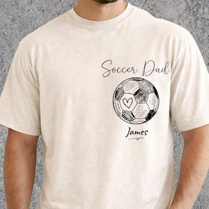 Może przedstawiać: Kremowa koszulka z czarno-białą grafiką piłki nożnej oraz napisami "Soccer Dad" i "James". Piłka nożna ma serce w środku. Koszulka jest wykonana z miękkiego materiału.