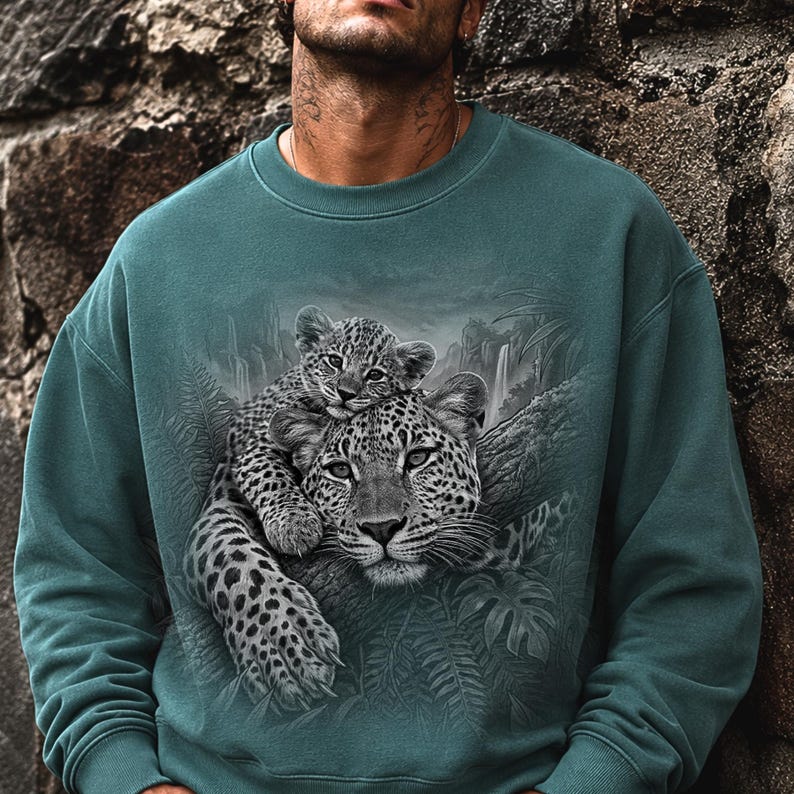 May include: A teal crewneck sweatshirt featuring a detailed graphic print of a leopard and its cub resting on a tree branch. The artwork includes lush jungle foliage and a waterfall in the background, rendered in shades of gray and black.