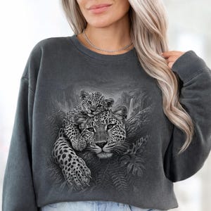 May include: A charcoal gray crewneck sweatshirt featuring a detailed black and white graphic print of a leopard resting on a tree branch with a cub perched on its back, surrounded by a jungle scene with ferns and a waterfall.
