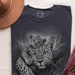 May include: A dark gray t-shirt featuring a graphic print of a leopard cub resting on an adult leopard in a jungle setting with waterfalls and ferns. The shirt has a Comfort Colors brand label at the neckline and is paired with a brown hat.
