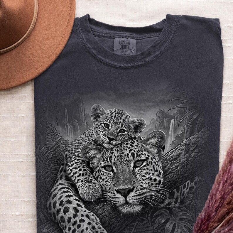 May include: A dark gray t-shirt featuring a graphic print of a leopard cub resting on an adult leopard in a jungle setting with waterfalls and ferns. The shirt has a Comfort Colors brand label at the neckline and is paired with a brown hat.