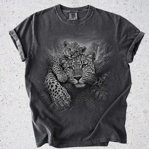 May include: A charcoal gray short-sleeve t-shirt featuring a detailed black and white graphic print of a leopard cub resting on the back of an adult leopard. The leopards are positioned on a tree branch surrounded by jungle foliage and a waterfall.