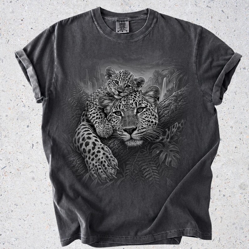May include: A charcoal gray short-sleeve t-shirt featuring a detailed black and white graphic print of a leopard cub resting on the back of an adult leopard. The leopards are positioned on a tree branch surrounded by jungle foliage and a waterfall.