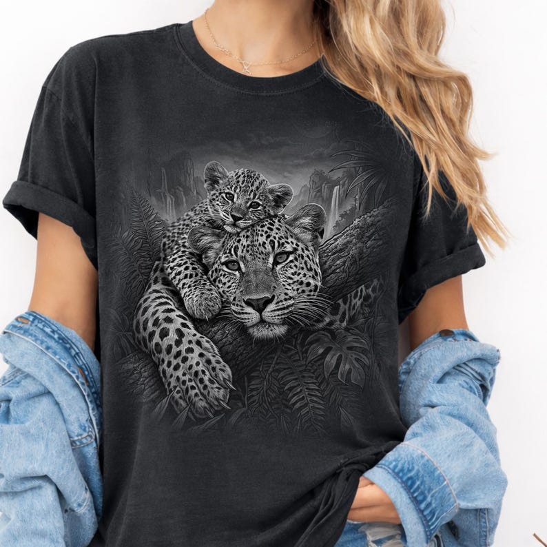 Comfort Colors® Pepper shirt featuring a detailed black and white leopard graphic with a leopard cub resting on top of an adult leopard in a jungle setting. Moody wildlife design with tropical leaves, rocky landscape, and realistic big cat artwork. Great animal graphic tee for leopard lovers, wildlife fans, safari style outfits, nature lovers, and unique animal gift ideas.