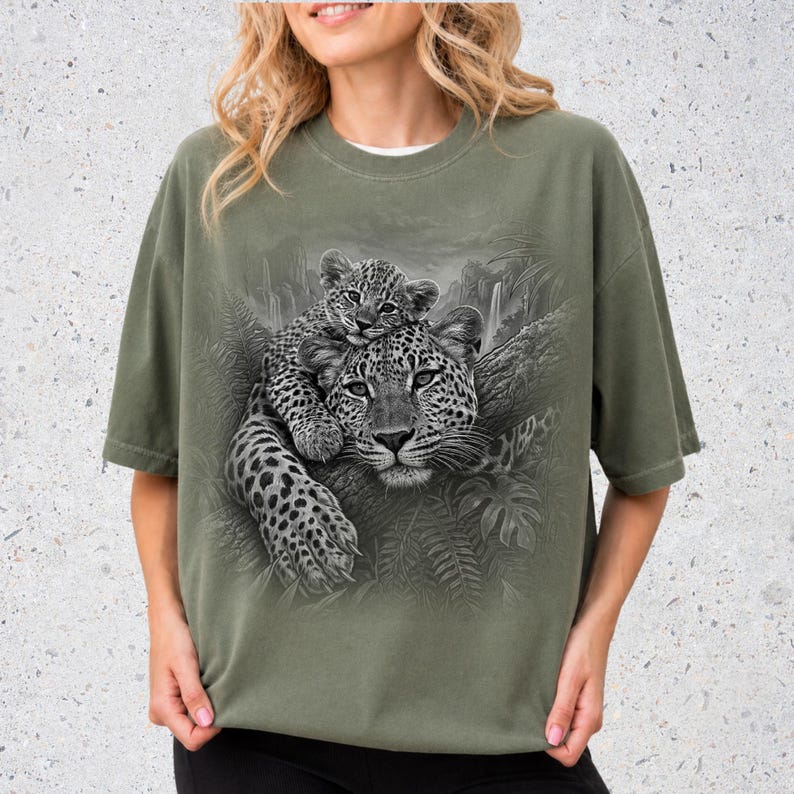 May include: An olive green short-sleeve t-shirt featuring a detailed black and white graphic print of a leopard resting on a tree branch with a cub perched on its back, surrounded by a jungle foliage and waterfall background.