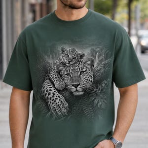 May include: A dark green short-sleeve t-shirt featuring a detailed monochromatic graphic print of a leopard resting on a tree branch with a cub perched on its back, set against a backdrop of lush jungle foliage and a waterfall.