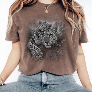 May include: A brown short-sleeve t-shirt featuring a detailed black and white graphic print of a leopard cub resting on the back of an adult leopard, surrounded by jungle foliage and trees in a natural, textured illustration style.
