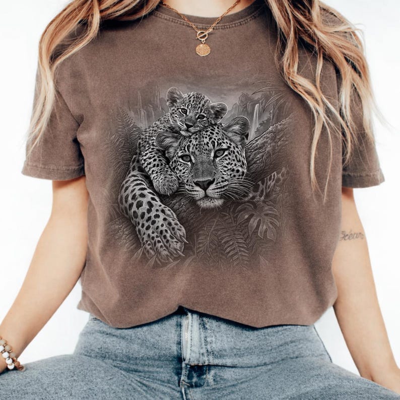 May include: A brown short-sleeve t-shirt featuring a detailed black and white graphic print of a leopard cub resting on the back of an adult leopard, surrounded by jungle foliage and trees in a natural, textured illustration style.