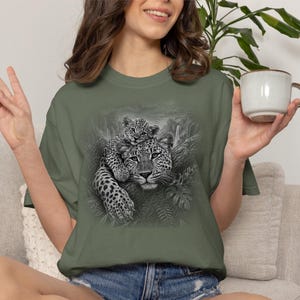May include: An olive green short-sleeve t-shirt featuring a detailed black and white graphic print of a leopard resting on a tree branch with a cub perched on its back, surrounded by lush jungle foliage and a waterfall in the background.