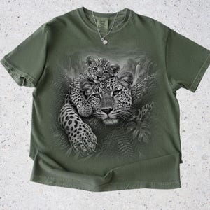 May include: An olive green short-sleeve t-shirt featuring a black and white graphic print of a leopard cub resting on an adult leopard in a jungle setting. The shirt has a crew neckline and a Comfort Colors brand label at the inner collar.