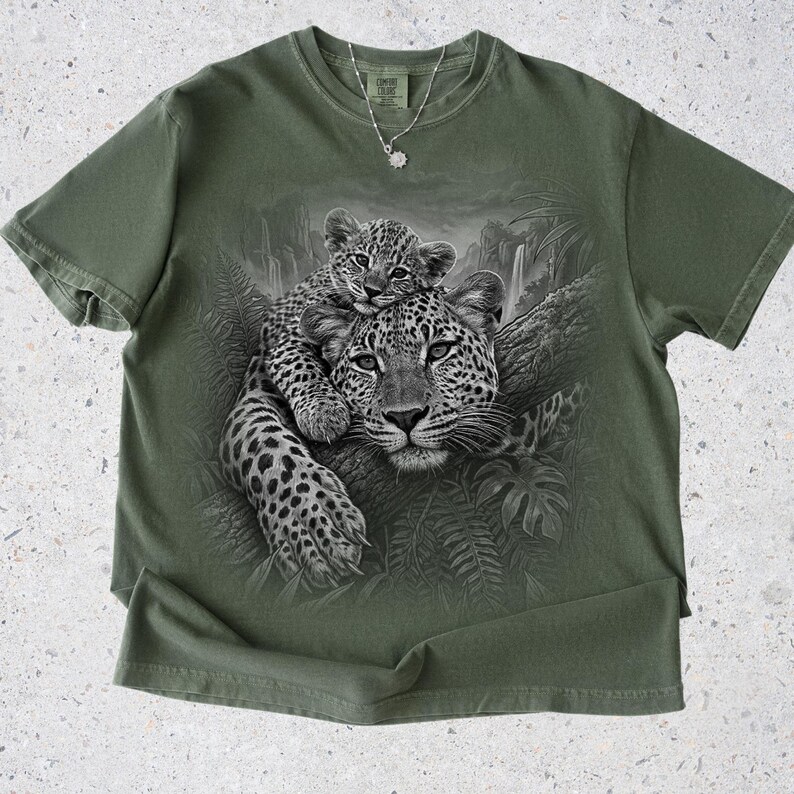 May include: An olive green short-sleeve t-shirt featuring a black and white graphic print of a leopard cub resting on an adult leopard in a jungle setting. The shirt has a crew neckline and a Comfort Colors brand label at the inner collar.