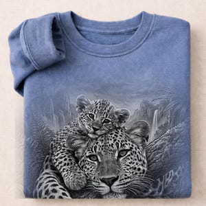 May include: A blue crew neck sweatshirt featuring a detailed graphic print of a leopard cub resting on top of an adult leopard. The background depicts a monochromatic jungle scene with waterfalls, foliage, and tree branches in shades of gray.