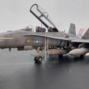 Op de afbeelding: Gedetailleerd model van een grijze militaire jet met open cockpit. Het model heeft een starembleem, raketten en een ladder. De jet staat op een donkere ondergrond. Gedetailleerde replica van een militair vliegtuig.