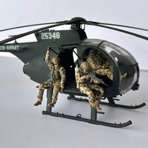 May include: A detailed model of a dark green military helicopter with the text "UNITED STATES ARMY" and the number "25348". Two miniature soldiers in camouflage uniforms are seated in the helicopter, and one is seated on the outside.