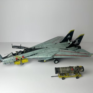 Può includere: Modello dettagliato di un caccia F-14 Tomcat grigio e azzurro con dettagli neri e gialli. Il modello include un motore giallo separato su un piccolo carrello. Il jet ha il numero 103 sul lato.