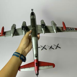 Pu&ograve; includere: Modello dettagliato di aereo con fusoliera argentata ed estremit&agrave; alari rosse. Le ali mostrano il testo "USAF" e un'emblema a stella. Quattro eliche nere sono posizionate vicino all'aereo.