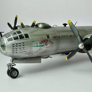 Pu&ograve; includere: Modello dettagliato di un aereo argentato con eliche nere e punte gialle. Le parole "UNITED STATES AIR FORCE" sono visibili sul lato. L'aereo presenta una grafica di ragazza pin-up e le parole "CARIBBEAN QUEEN".
