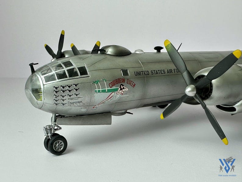 Pu&ograve; includere: Modello dettagliato di un aereo argentato con eliche nere e punte gialle. Le parole "UNITED STATES AIR FORCE" sono visibili sul lato. L'aereo presenta una grafica di ragazza pin-up e le parole "CARIBBEAN QUEEN".