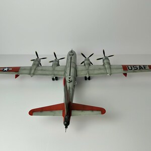Pu&ograve; includere: Un modello dettagliato di aereo, con una combinazione di colori argento e rosso e quattro eliche. Le ali mostrano il testo "USAF" e un'emblema a stella. Il modello &egrave; una replica di un aereo d'epoca.