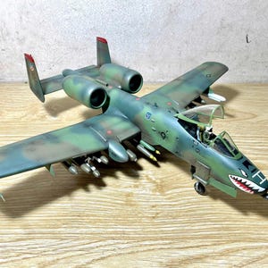 May include: A detailed model of a military aircraft, painted in camouflage green and brown. The model features a shark mouth design on the nose and is equipped with missiles. The wings and tail have red accents. The model is a replica of the A-10 Thunderbolt II.