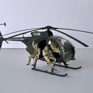 May include: A dark green model helicopter with the text "UNITED STATES ARMY" and the number "25348". Three figures in camouflage uniforms are positioned near the open door. The helicopter has a clear cockpit and a black rotor.