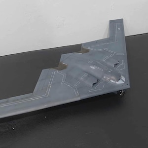 May include: A grey model of a B-2 Spirit stealth bomber aircraft. The model has a distinctive flying wing design with a dark grey colour scheme and detailed panel lines. The aircraft is resting on a black surface.
