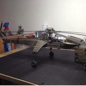 Pode incluir: Modelo detalhado de um helicóptero militar, principalmente verde-oliva, com um complexo sistema de rotor e trem de aterrissagem. O modelo está sobre uma superfície de grade preta, mostrando o design.