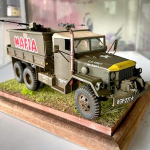 Può includere: Modello dettagliato di un camion militare verde oliva con frontale giallo. Il camion presenta la scritta "THE MAFIA" in rosso sul lato ed è montato su una base di legno con terreno erboso.