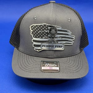 May include: A grey and black trucker hat featuring a patch with a distressed American flag, a skull wearing a welding helmet, and the text PUDDLE PIMP. The brim has a sticker with the text BrimCo EST 2008 authentic 100 trucker.