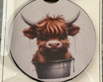 May include: Round car coaster featuring a Highland cow illustration. The cow has brown fur, large horns, and is depicted in a bucket. The coaster has a black border and a light gray background.