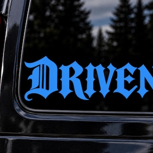 May include: A bright blue vinyl decal applied to a vehicle window, featuring the word Driven written in a bold, stylized gothic font with sharp, pointed edges and intricate letterforms that stand out against the dark glass surface.