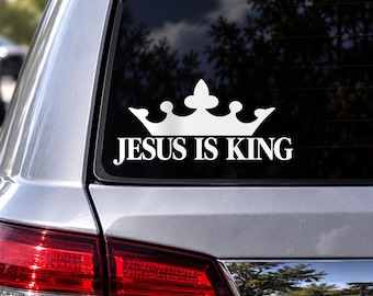 Puede incluir: Un adhesivo de vinilo blanco aplicado en la luneta trasera de un coche, con un gráfico de corona estilizada sobre el texto "JESUS IS KING" en una fuente serif negrita. El diseño es monocromático y visible sobre el cristal oscuro.