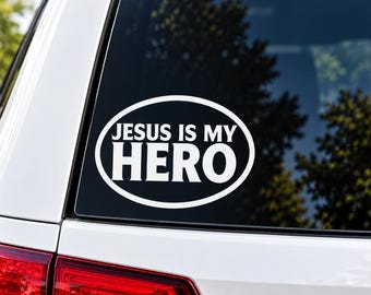 Puede incluir: Una pegatina de vinilo ovalada de color blanco aplicada en la luneta trasera de un vehículo. La pegatina presenta un borde blanco grueso y el texto "JESUS IS MY HERO" en una fuente sans-serif, con la palabra "HERO" en un tamaño mayor al resto.