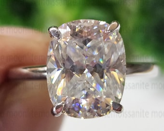May include: A close-up of a silver ring featuring a large, cushion-cut, light-colored gemstone. The gemstone is held in place by four prongs. The background is blurred, with green foliage visible.