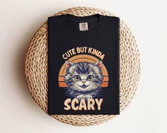 May include: Black t-shirt with a cartoon cat design and the words "Cute but Kinda Scary." The cat has grey and white fur, orange eyes, and is set against an orange and yellow sunset. The shirt is folded on a woven, circular surface.