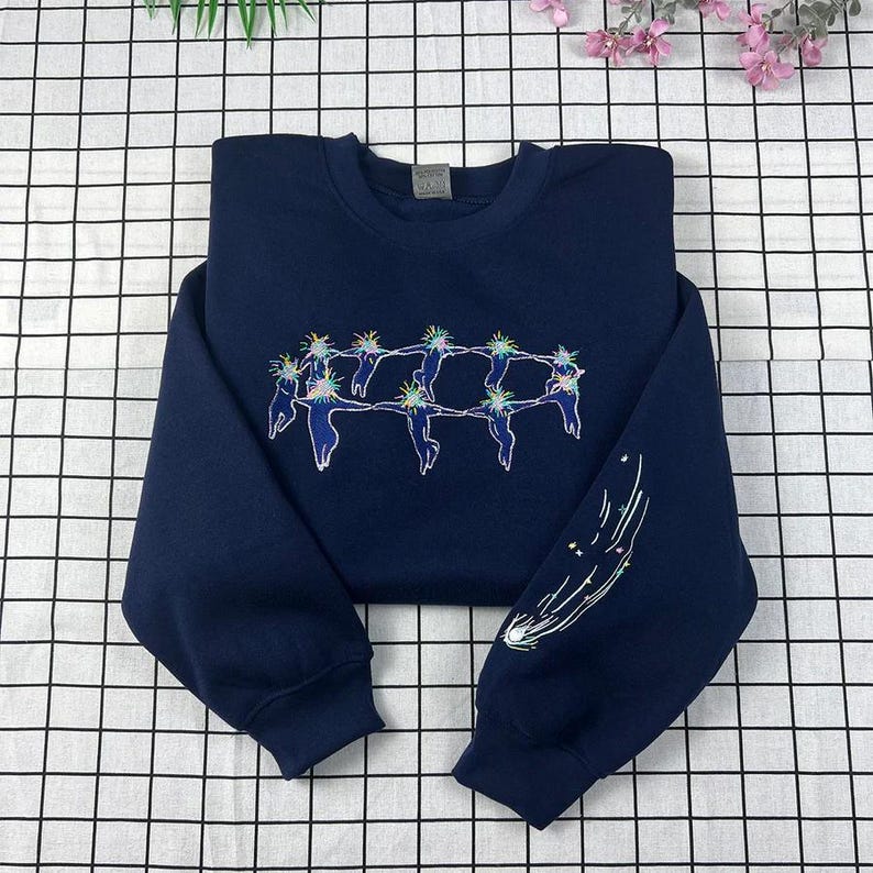 May include: Navy blue sweatshirt with embroidered celestial design. The design features a circle of figures with star-like elements above their heads, and a comet on the sleeve. The sweatshirt is made of a soft material.