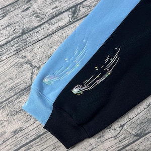 May include: Two long-sleeved sweatshirts, one light blue and one black, are displayed. Each sleeve features an embroidered comet design with colorful stars and a white trail. The cuffs and lower hems are solid colors.