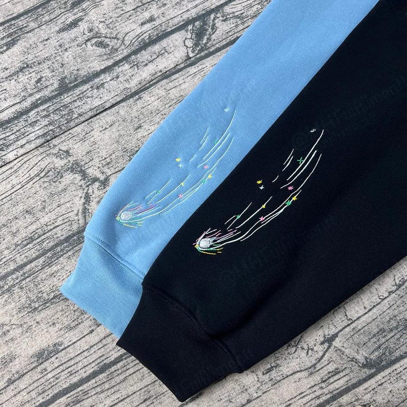 May include: Two long-sleeved sweatshirts, one light blue and one black, are displayed. Each sleeve features an embroidered comet design with colorful stars and a white trail. The cuffs and lower hems are solid colors.