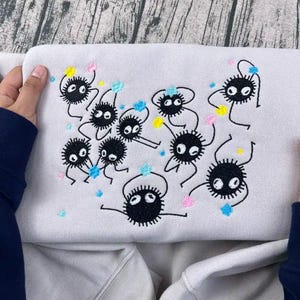 Op de afbeelding: Een lichtgrijs sweatshirt met een geborduurd ontwerp van verschillende zwarte, ronde, cartoonachtige wezens met witte ogen en stekelige lichamen. Het ontwerp bevat kleurrijke accenten van roze, blauw en geel.