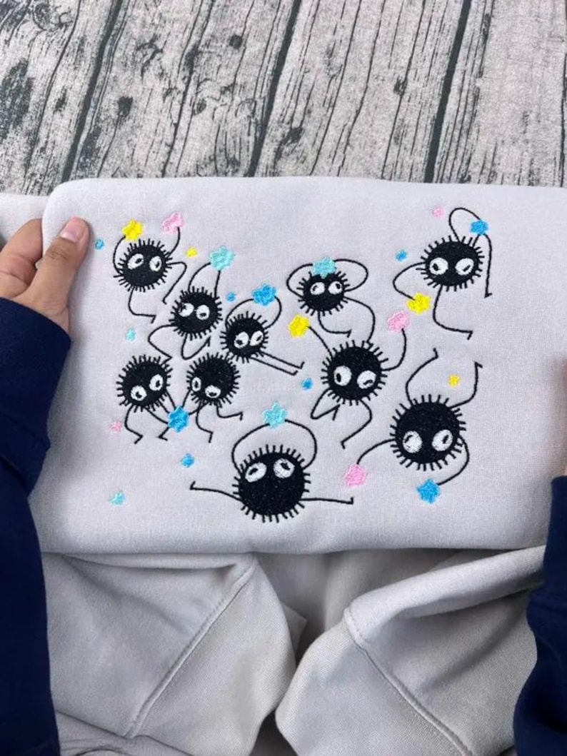 May include: A light gray sweatshirt featuring an embroidered design of several black, round, cartoon-like creatures with white eyes and spiky bodies. The design includes colorful accents of pink, blue, and yellow.