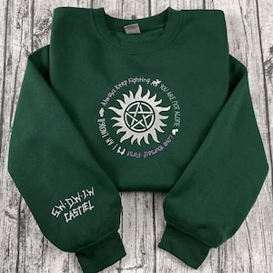 Könnte beinhalten: Ein dunkelgrünes Sweatshirt mit weißem und lila gesticktem Text und Design. Das Design umfasst einen Sonnenaufgang mit einem Pentagramm in der Mitte, umgeben von den Sätzen "Always Keep Fighting", "You Are Not Alone", "Love Yourself First" und "I Am Enough". Der linke Ärmel hat den gestickten Text "S.W. D.W. J.W. CASTIEL".