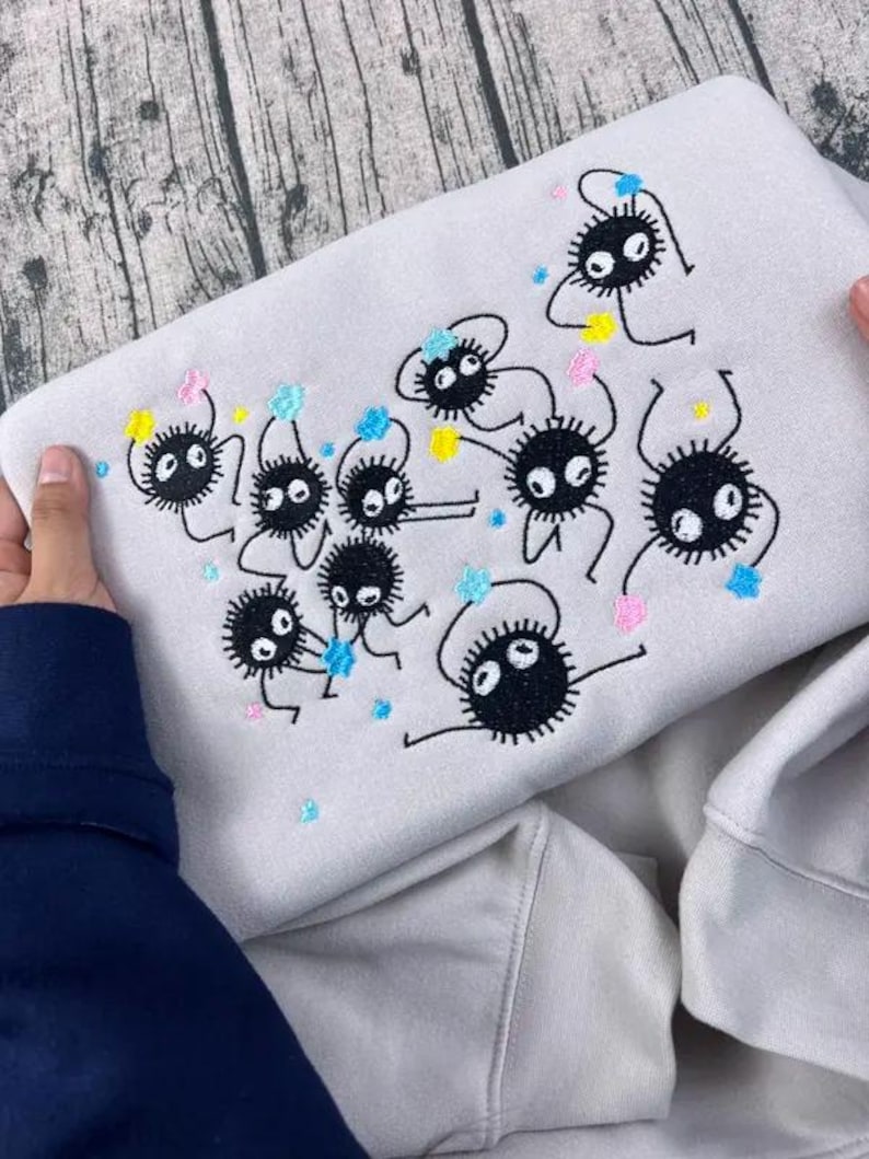May include: A light gray sweatshirt featuring an embroidered design of black, round, cartoon-like creatures with large eyes and small limbs. The design includes colorful accents of blue, pink, and yellow. The sweatshirt is held by hands.