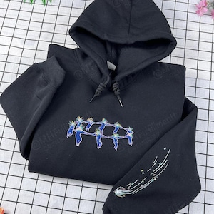 May include: Black hooded sweatshirt with a drawstring hood. The front features a colorful embroidered design of figures in a circle. The sleeve has a white and colorful shooting star design. The sweatshirt is made of a soft material.