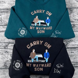 Może przedstawiać: Dwie bluzy, jedna morska i jedna czarna, z haftowanymi wzorami. Bluza morska ma napis "CARRY ON" nad wizerunkiem dwóch postaci i samochodu, z napisem "MY WAYWARD SON" poniżej. Czarna bluza ma ten sam wzór. Wymiary w cm.