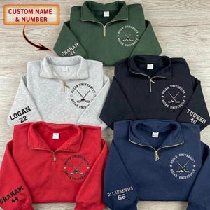 Op de afbeelding: Een verzameling quarter-zip truien in groen, grijs, zwart, rood en marineblauw. Elke trui heeft een "Briar University" logo en kan worden gepersonaliseerd met een naam en nummer. De tekst "CUSTOM NAME & NUMBER" is ook zichtbaar.