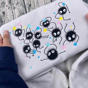 May include: A light gray sweatshirt featuring an embroidered design of several black, round, cartoon-like creatures with large eyes and small limbs. The design includes colorful accents of blue, yellow, and pink. The sweatshirt is held by two hands.