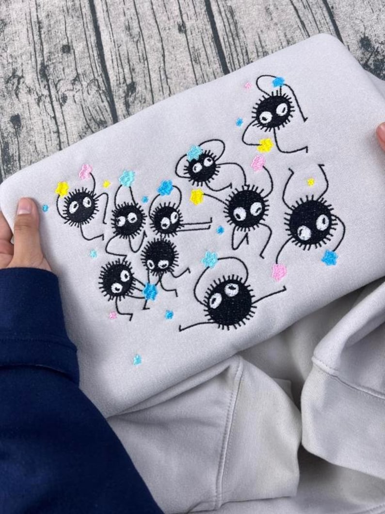 May include: A light gray sweatshirt featuring an embroidered design of several black, round, cartoon-like creatures with large eyes and small limbs. The design includes colorful accents of blue, yellow, and pink. The sweatshirt is held by two hands.