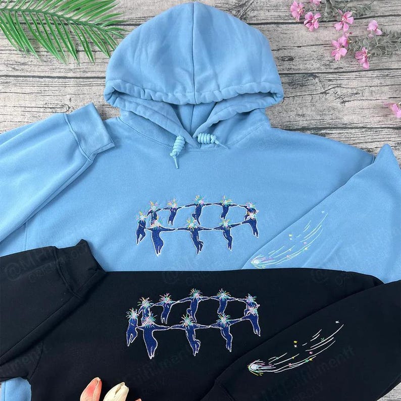 May include: Light blue hoodie with a drawstring hood and embroidered design of figures in a circle. The sleeve features a shooting star design. A black hoodie with the same design is also visible.