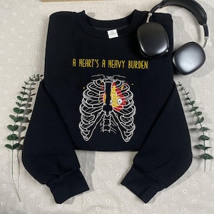 Op de afbeelding: Zwarte crewneck sweatshirt met de gouden tekst "A Heart's A Heavy Burden". De afbeelding toont een witte ribbenkast met een rood en geel hartontwerp. Zwarte koptelefoon op de achtergrond.