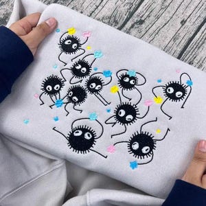 May include: A light gray sweatshirt featuring an embroidered design of several black soot sprites with white eyes and colorful accents. The design includes yellow, pink, and blue details. The sweatshirt is held by a person.